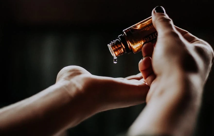A person pours liquid from a small amber bottle onto their hand against a dark, blurred background, suggesting a step in their detox routine.