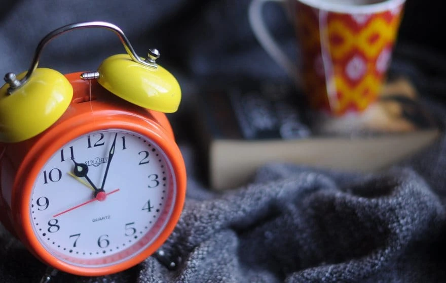 An orange and yellow analogue alarm clock shows 8:00, placed on a grey fabric with a book and a red patterned mug in the blurred background—perfect for pondering the best time for your vitamin IV drip.