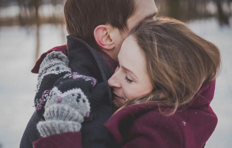 A woman and a man embrace outdoors in winter clothing, with the woman’s eyes closed and knitted mittens visible. Snow and trees are in the background, capturing a moment of winter wellbeing in the crisp, fresh air.