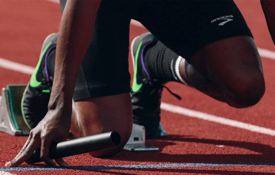 A runner kneels in the starting blocks on a track, holding a relay baton and wearing running shoes and sports kit, ready to enhance performance.