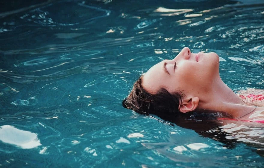 A person floats on their back in a swimming pool with eyes closed, surrounded by clear blue water, enjoying a sense of cleansing and renewal.