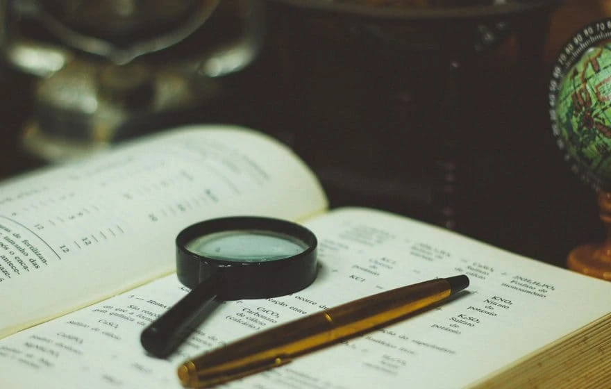A magnifying glass and a pen rest on an open book filled with printed text, suggesting study or research activity—perhaps delving into the history of intravenous therapy or its development through the years.