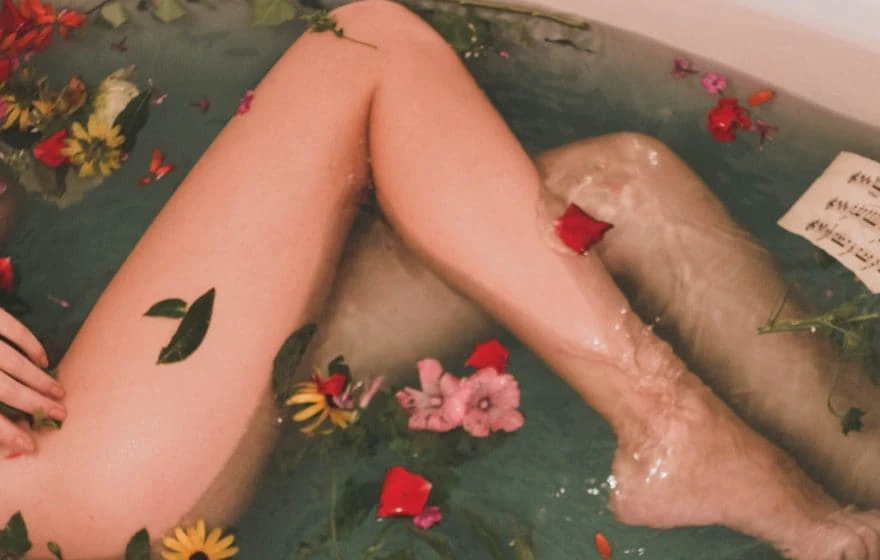 Person’s legs in a bath filled with water, colourful flower petals, and leaves floating on the surface—creating a serene moment that highlights glowing skin.