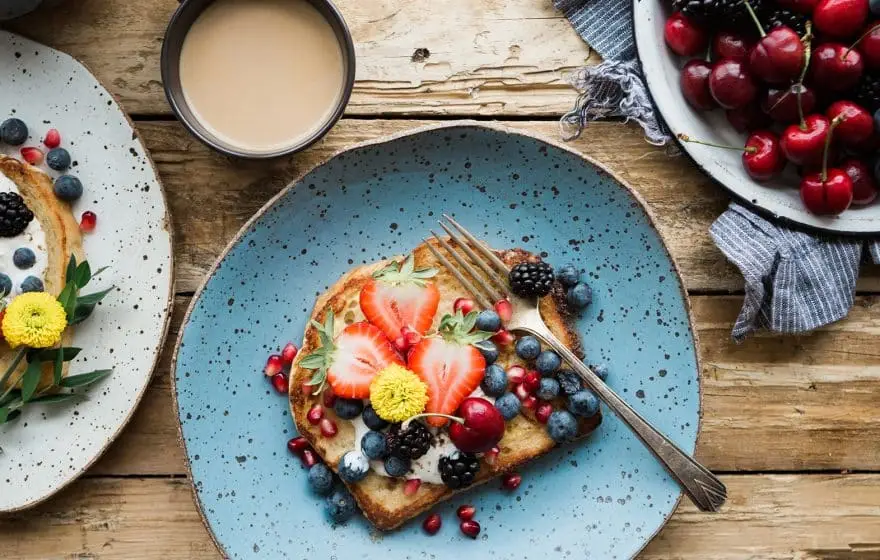 A plate with toast topped with yoghurt, superfoods like strawberries, blueberries, blackberries, pomegranate seeds, and a yellow edible flower sits beside a bowl of cherries and a cup of coffee—perfect to supercharge your healthy new year resolutions.