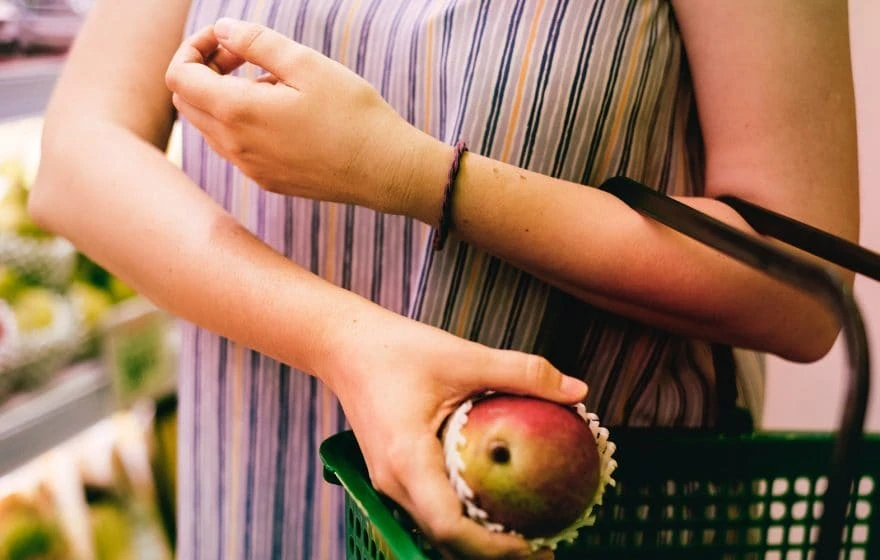 A person in a striped shirt holds a green shopping basket and picks up a mango in a supermarket, mindful of hidden risks that certain foods may pose for those on a restricted diet.