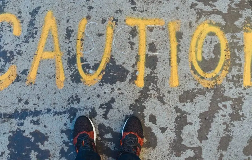 Yellow CAUTION is painted on worn pavement, partially seen from above, with a person's feet in trainers visible at the bottom—echoing caution around life expectancy trends similar to recent UK life expectancy concerns.