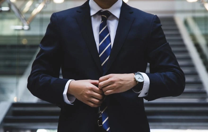 Person in a dark suit and striped tie fastens their jacket while standing in front of a staircase, exuding a professional edge.