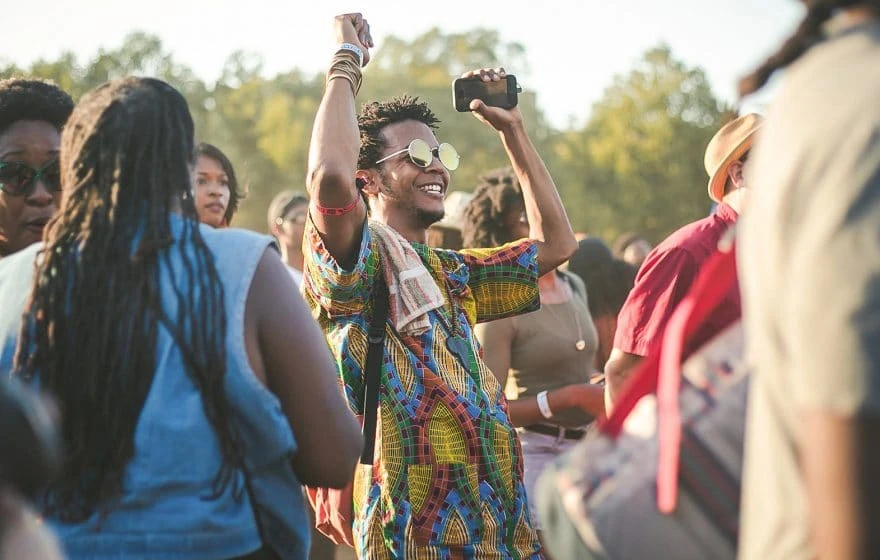 A person wearing colourful clothing and sunglasses raises their hands whilst dancing in a crowd at a summer festival. Trees are visible in the background, capturing the excitement of festival season.