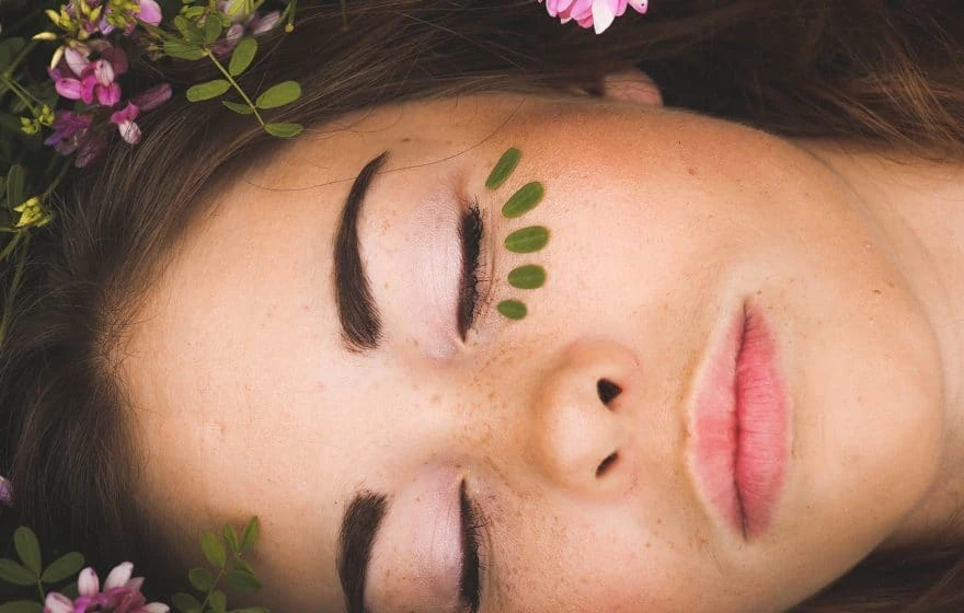 A close-up of a person with closed eyes, freckles, and green leaf petals under one eye. Pink flowers and green leaves frame their face, highlighting the natural beauty of beautiful skin.
