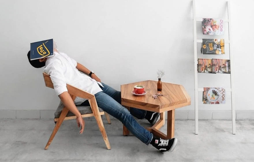 A person sits slouched in a wooden chair with a book covering their face, next to a small table holding a drink and a vase, beside a magazine rack against a white wall.
