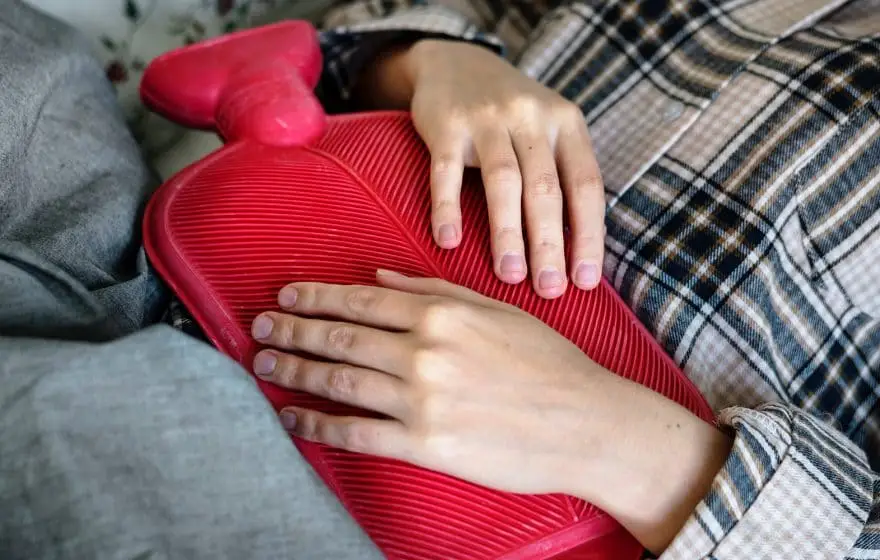 A person lying down holds a red rubber hot water bottle against their stomach with both hands, highlighting common misconceptions about managing Lyme Disease symptoms.