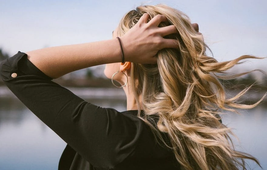 A person with long, wavy blonde hair stands outdoors near water, facing away and running a hand through their hair—a gentle reminder that prioritising hair care could be the perfect New Year’s Resolution. They wear a black top and a hoop earring.