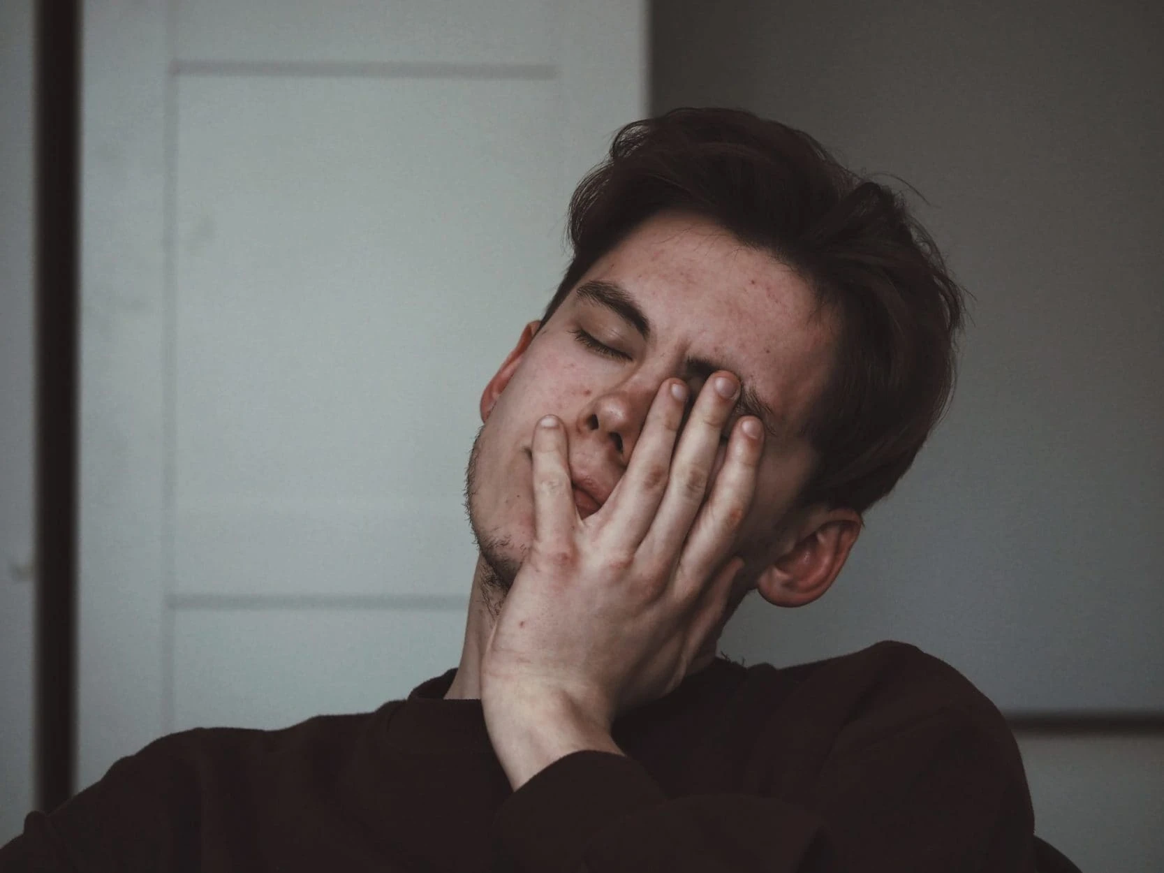 A person with short brown hair rests their face on their hand, eyes closed, appearing tired or stressed—common symptoms of Vitamin B12 deficiency—in an indoor setting with neutral-coloured walls.