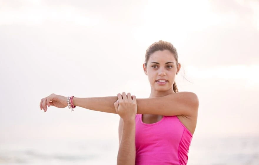 A woman in a pink vest top stretches her arm across her chest outdoors, looking towards the camera—embracing wellbeing much like exploring the 7 Major Benefits of NAD+ IV Therapy.