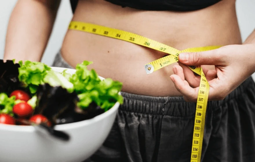 A person measures their waist with a tape measure whilst holding a bowl of salad with lettuce and tomatoes, highlighting their commitment to weight loss.