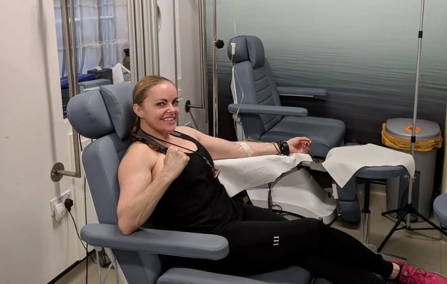 A woman sits in a medical chair at IV Boost Clinic, donating blood or plasma, smiling and flexing her arm with a drip line attached.