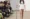 A model walks down a catwalk wearing a white T-shirt with the word SICK printed on it, holding a drip stand—a striking nod to health and IV treatments—while members of the audience watch and take photos.