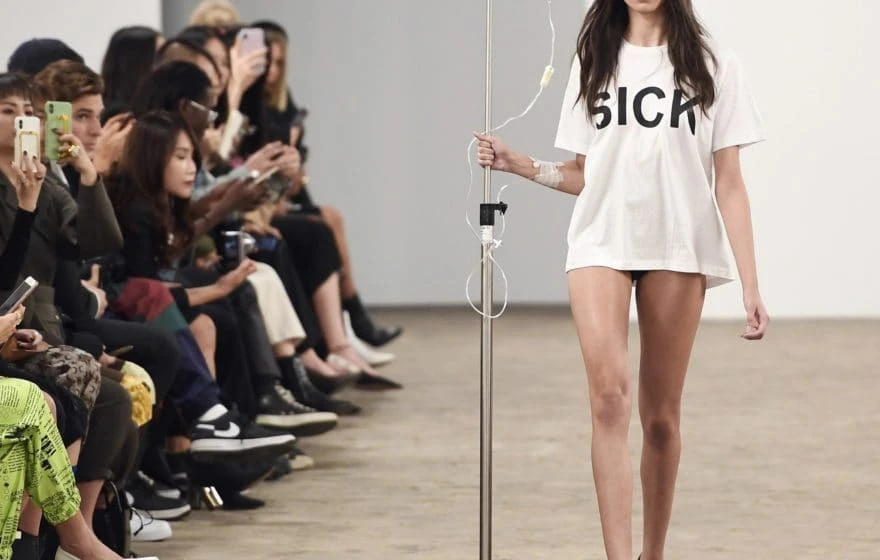 A model walks down a catwalk wearing a white T-shirt with the word SICK printed on it, holding a drip stand—a striking nod to health and IV treatments—while members of the audience watch and take photos.