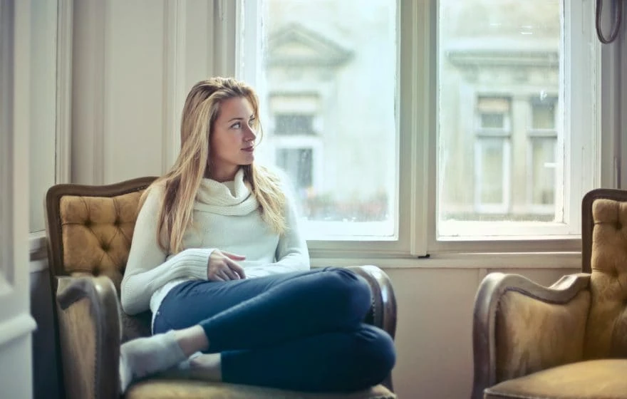 A woman in a white jumper and blue jeans sits cross-legged on an armchair, looking out of a window during lockdown in a well-lit room with vintage decor.
