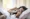 A woman lies in bed under a white blanket, holding a tissue to her nose as if sneezing or wiping it, perhaps following Lifestyle tips from Dr Joshua Berkowitz for recovery from illness.