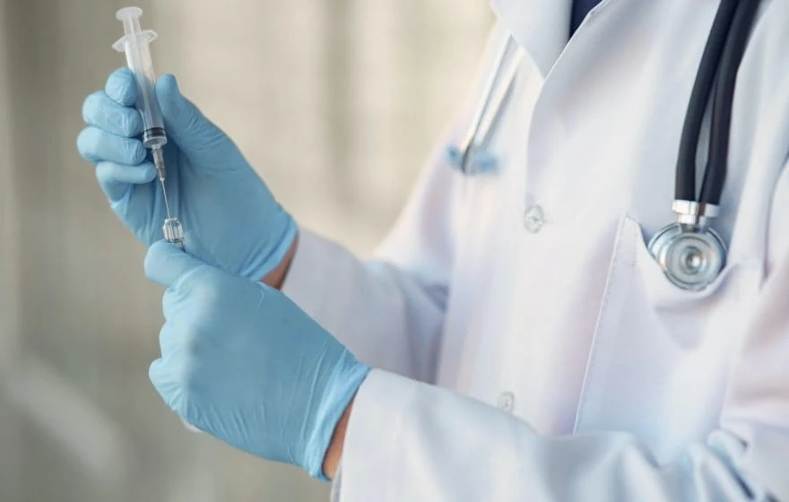 A healthcare professional wearing a white coat and blue gloves prepares a syringe with liquid Vitamin B12, with a stethoscope draped around their neck.