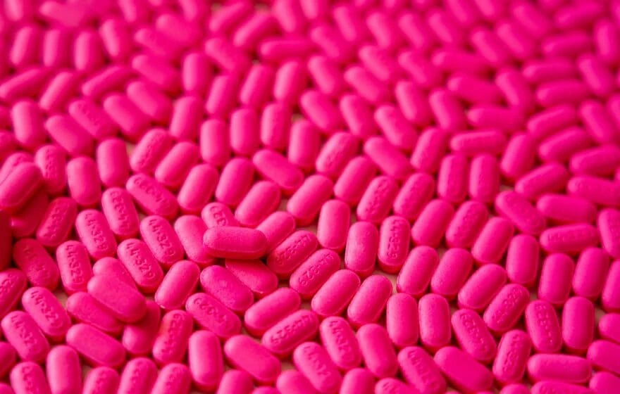 A large number of bright pink, oval-shaped tablets—possibly supplements such as Vitamin B12 vs B Complex—are spread out across a surface.