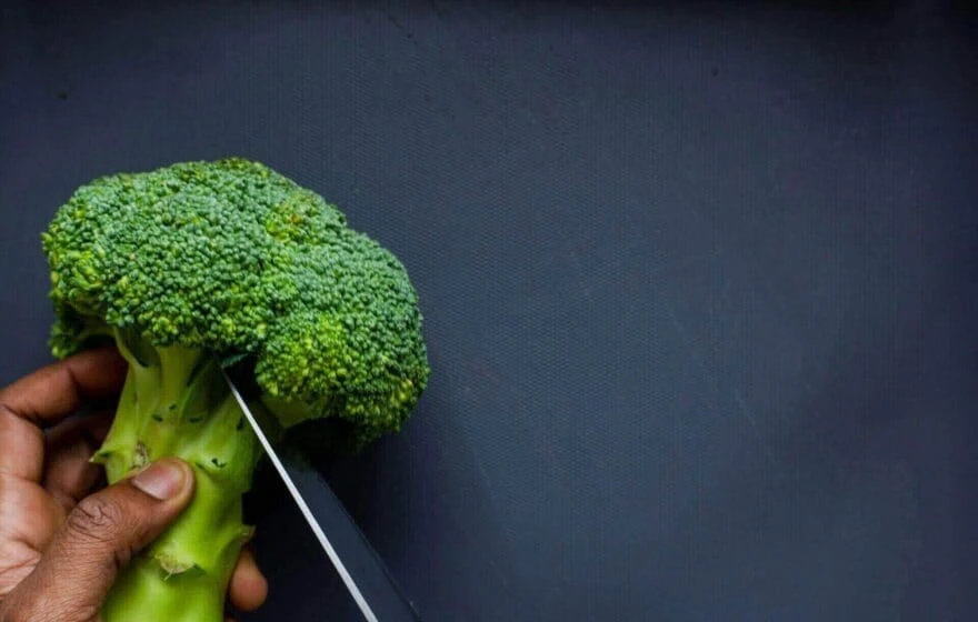 A hand holds a head of broccoli while a knife slices through the stem against a dark background—a nod to healthy eating and natural sources of antioxidants like glutathione.