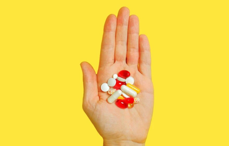 A hand holding various pills and capsules against a solid yellow background, illustrating how vitamin absorption works.