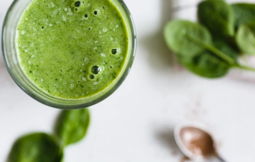 A close-up of a glass filled with a green smoothie, with fresh spinach leaves and a spoon with powder in the background—perfect for those seeking tips for better nutrition.