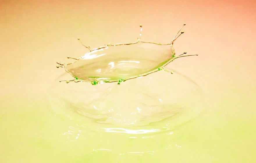 A high-speed photo captures a water droplet splashing, forming a crown-like shape against a soft pink and green gradient background—evoking the playful colours of a banana bag.