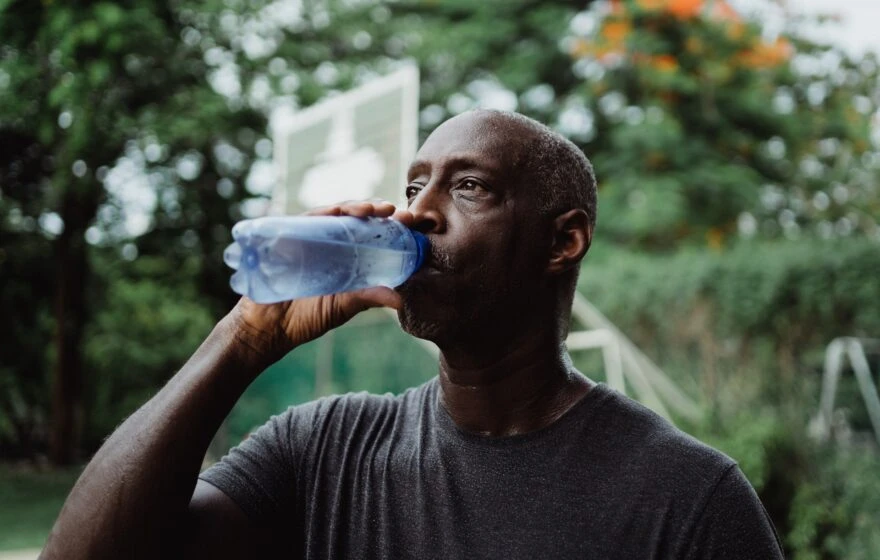 A man in a dark T-shirt drinks water from a blue bottle outdoors, staying hydrated to help combat dehydration and anxiety, with greenery and a basketball hoop blurred in the background.
