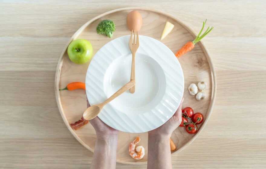 A person holds a white plate with wooden utensils arranged like clock hands, symbolising ways to boost metabolism; the plate rests on a tray with fruit and vegetables arranged in a circle.