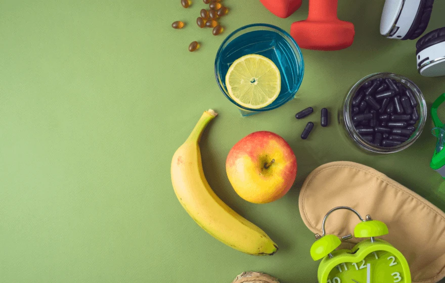 Banana, apple, a glass of water with lemon, dumbbells, supplements, headphones, sleep mask, and alarm clock arranged on a green surface—a perfect snapshot of biohacking essentials for a healthier lifestyle.