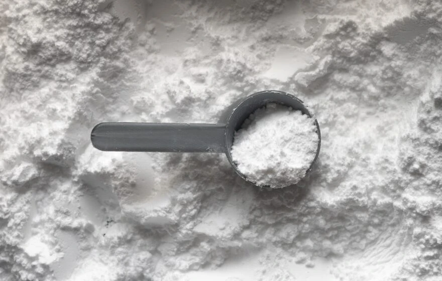 A grey plastic scoop filled with white powder, likely a glutamine supplement, rests on a surface covered in the same powder.