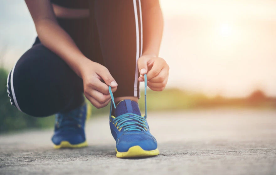Kneeling on the ground and tying the shoelaces of a blue trainer, a person in black trousers with white stripes prepares for peak athletic performance outdoors in natural light.