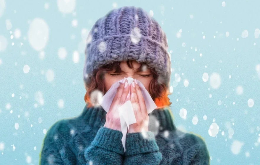 A person wearing a knitted hat and jumper blows their nose with a tissue while standing in falling snow, highlighting the importance of winter health and ways to boost immunity during the colder months.