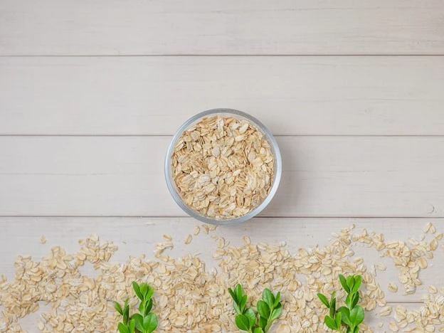 A bowl of porridge oats sits on a light wooden surface, with scattered oats and green leaves arranged at the bottom—highlighting the health benefits and cysteine content found in this nutritious breakfast choice.