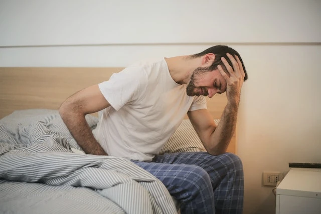 A man in pyjamas sits on the edge of a bed, holding his head with one hand and looking down, appearing unwell—perhaps in need of an instant relief hangover cure.