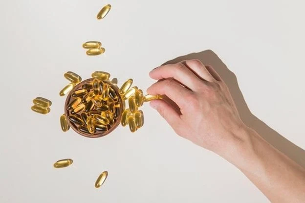 A hand reaching for a yellow Vitamin B12 capsule from a small bowl filled with similar capsules, highlighting the importance of health, with some capsules scattered on a white surface.