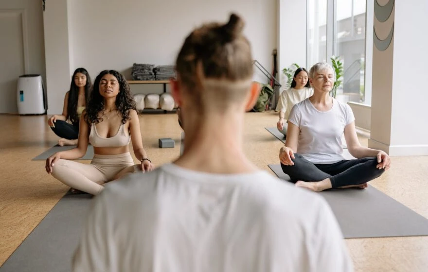 A group of people sit cross-legged on yoga mats in a bright room, meditating with eyes closed, embracing healthy ageing as they follow an instructor in the foreground.