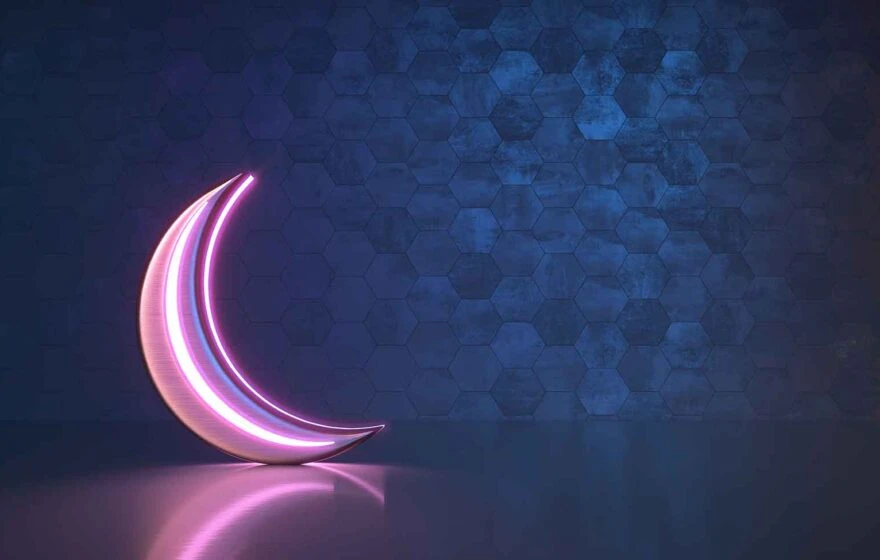 A pink neon crescent moon glows against a blue hexagonal-patterned wall, reflecting softly on a smooth surface—perfect inspiration for Ramadan fasting tips to help you ease out during the holy month.