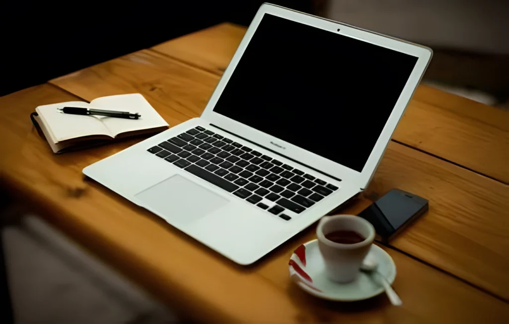 A silver laptop sits on a wooden table next to a notebook with a pen, a mobile phone, and a cup of tea on a saucer—an inviting space for researching topics like How Hydrogen Peroxide IV Therapy Works.