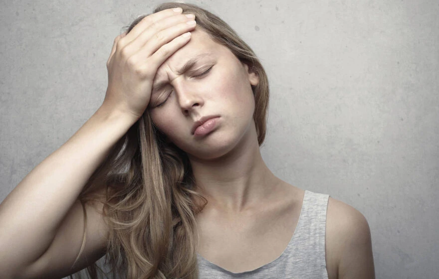 A young woman with long hair holds her hand to her forehead and closes her eyes, appearing to be in discomfort or pain—possibly in need of Vitamin B12 injections.