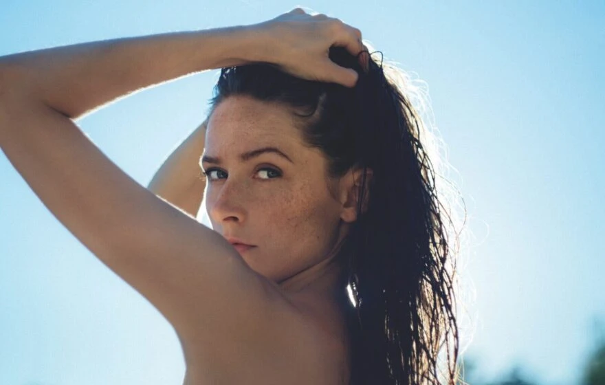 A woman with wet hair holds it back with one hand, looking over her shoulder towards the camera against a bright, sunlit background—a reminder of the importance of sunlight in preventing Vitamin D deficiency.