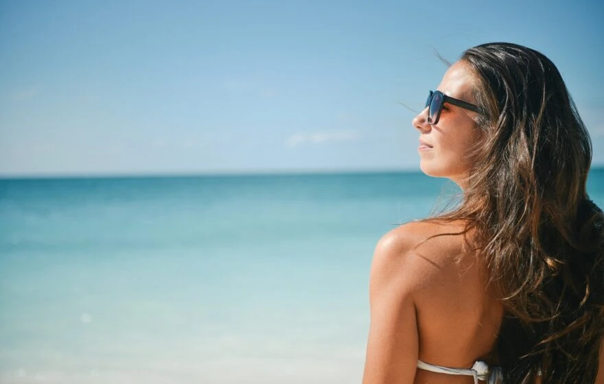 A woman wearing sunglasses and a bikini stands on a beach, soaking up Vitamin D2 and D3 as she gazes at the calm, blue sea under a clear sky.