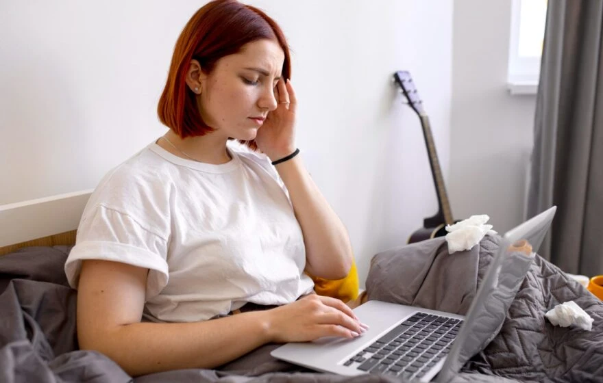 A woman with short red hair sits in bed using a laptop, looking unwell and touching her head, possibly due to Vitamin B12 deficiency, with tissues nearby and a guitar in the background.