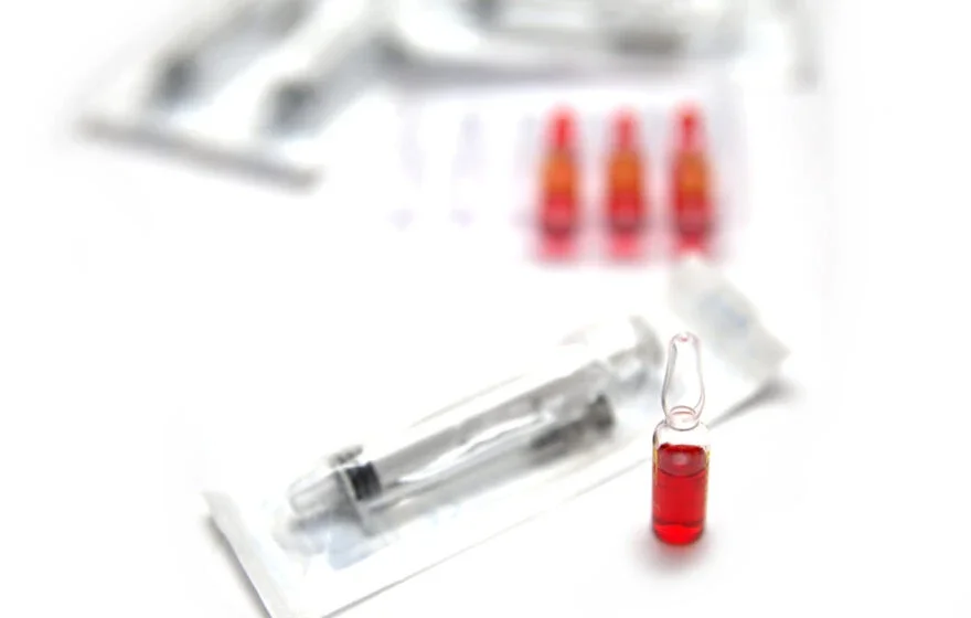 A glass ampoule filled with red liquid stands beside a packaged syringe, hinting at discussions on Vitamin B12 Injections Dosage and Frequency, with more ampoules and syringes blurred in the background.