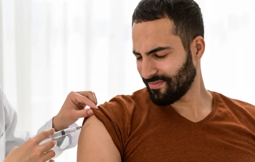A person in a brown shirt receives a Vitamin D injection in the upper arm from a healthcare professional.