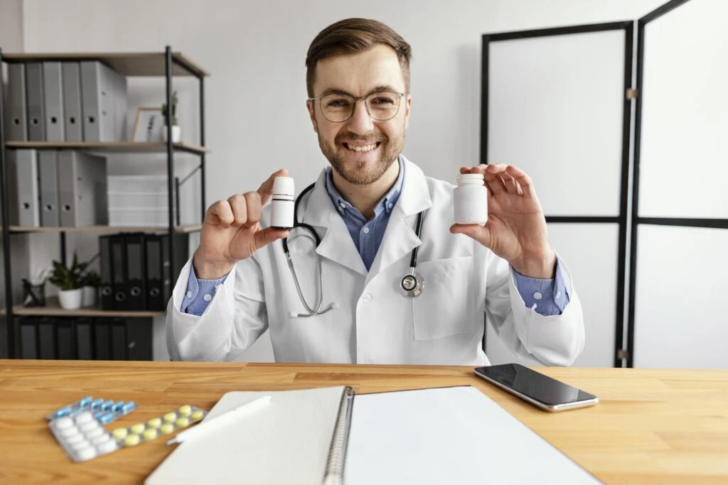 A male doctor wearing a white coat and stethoscope sits at a desk, smiling and holding two medicine bottles—one labelled vitamin B12 injection—with a notebook, mobile, and tablets on the table.