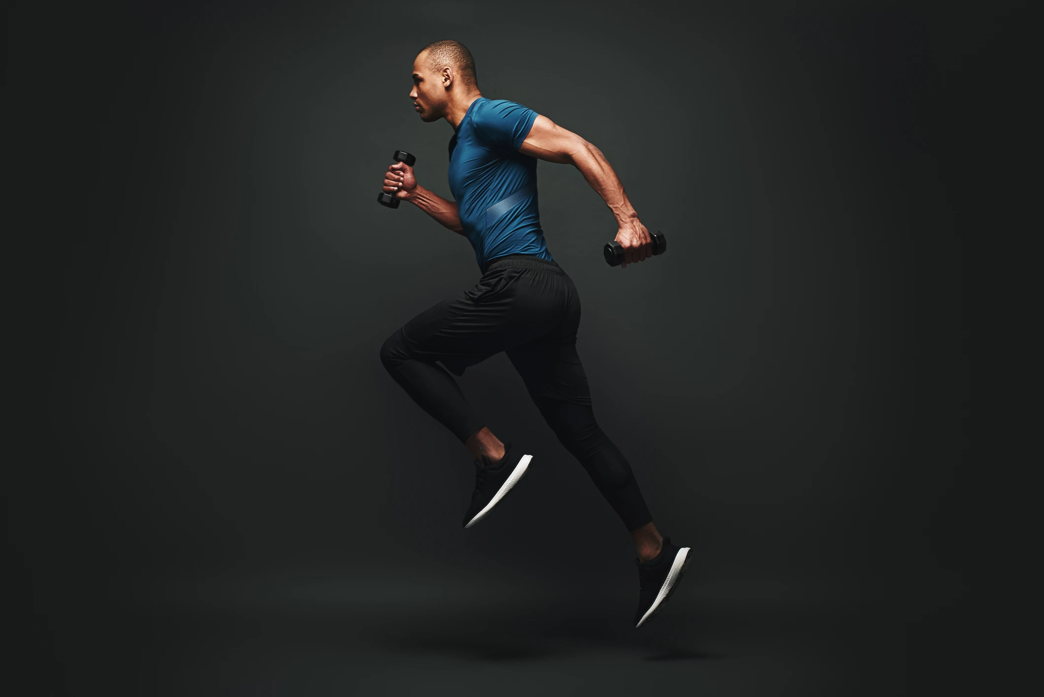 Man in athletic wear runs on the spot while holding a dumbbell in each hand against a dark background, embodying peak fitness—discover how IV Therapy can help you perform like an athlete and reach your own health goals.